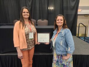 Two women standing in front of a stage, smiling at the camera. One woman holds a framed certificate that reads Ozark Center. Both are wearing conference badges around their necks.
