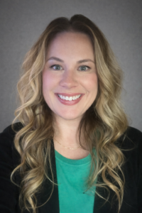 A woman with long, wavy blonde hair smiles at the camera. She is wearing a green top, a black jacket, and a thin gold necklace, with a neutral gray background behind her.