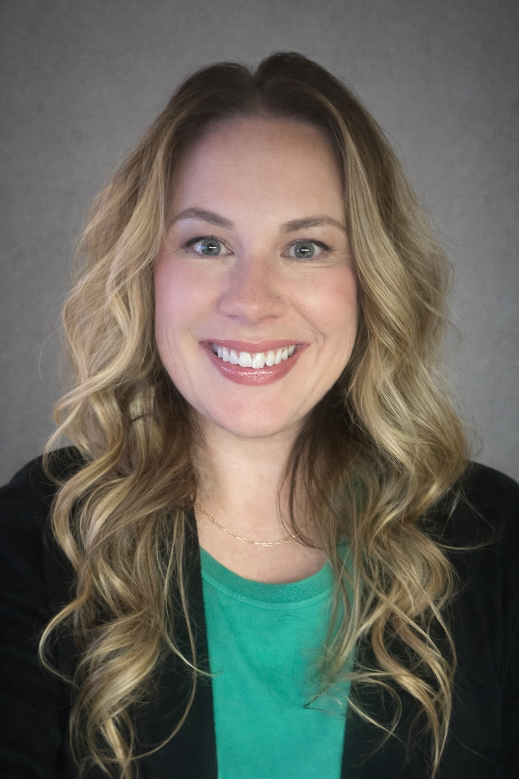 A woman with long, wavy blonde hair smiles at the camera. She is wearing a green top, a black jacket, and a thin gold necklace, with a neutral gray background behind her.