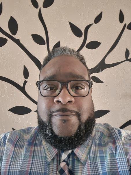 A man with glasses, a beard, and a mustache smiles at the camera. He is wearing a plaid shirt and tie. Behind him, a wall decoration with black branches and leaves appears to be sprouting from his head.