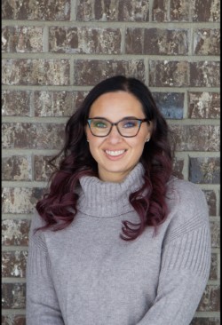 A woman with long, dark, wavy hair and glasses smiles at the camera. She is wearing a light gray turtleneck sweater and standing in front of a brick wall background.