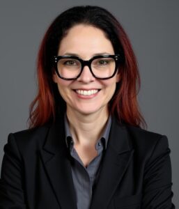 A woman with long dark hair and red highlights, wearing large black glasses, a dark blazer, and a gray shirt, smiles in front of a plain gray background.