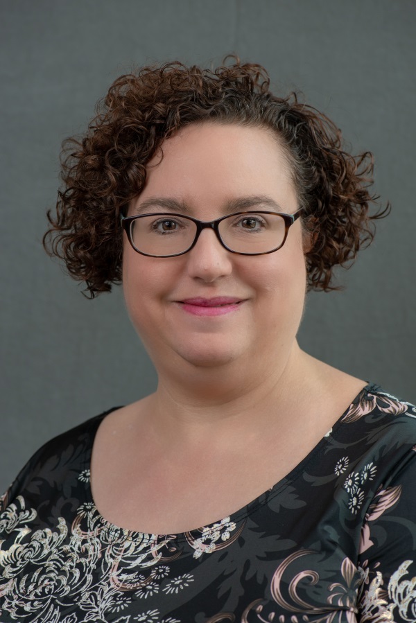 A woman with short curly brown hair and glasses smiles at the camera. She is wearing a black top with a floral pattern, and the background is plain gray.
