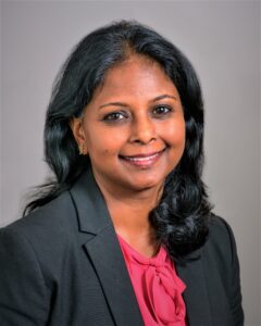 A woman with medium skin tone and long black hair, wearing a black blazer and a pink blouse, is smiling at the camera against a plain light-colored background.