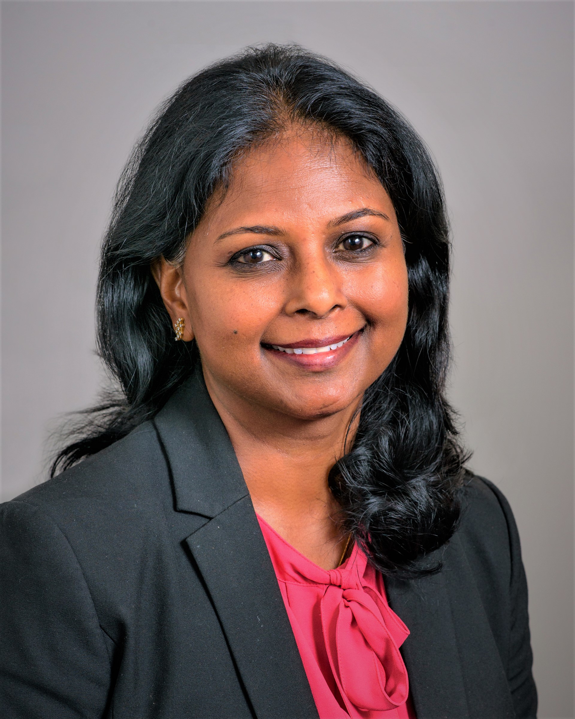 A woman with medium skin tone and long black hair, wearing a black blazer and a pink blouse, is smiling at the camera against a plain light-colored background.