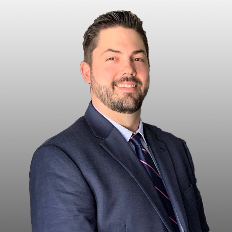 A man with short dark hair and a beard, wearing a navy blue suit, striped tie, and light shirt, smiles at the camera against a plain light gray background.