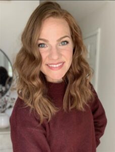 A woman with wavy, shoulder-length auburn hair and blue eyes smiles at the camera. She is wearing a maroon sweater and standing indoors in a softly lit room.
