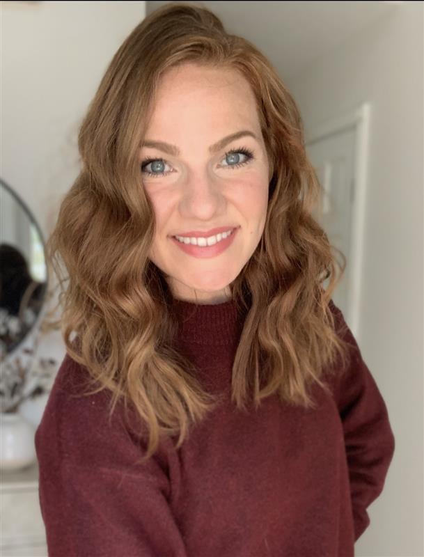 A woman with wavy, shoulder-length auburn hair and blue eyes smiles at the camera. She is wearing a maroon sweater and standing indoors in a softly lit room.
